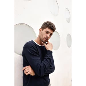 A model in a navy round-neck pullover stands against a textured wall with circular cutouts. The sweater features a white collar.
