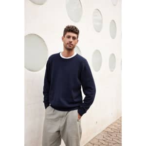A man wearing a navy round-neck pullover with a white collar, posing in front of a textured white wall.