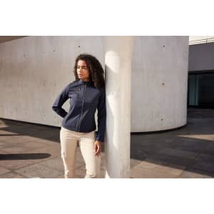 A navy ladies softshell jacket is worn by a model leaning against a column in an urban setting.
