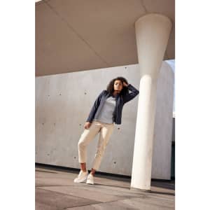 A woman wearing a navy softshell jacket, grey top, and cream pants, leaning against a column in an urban setting.