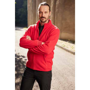 A man wearing a red bonded fleece jacket stands with arms crossed outdoors. The background features a stone wall.