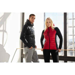 A hybrid jacket featuring black and red sections, worn by a man and woman standing indoors. The design includes a hood.
