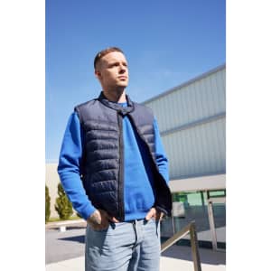 A padded navy vest worn over a blue jumper, modelled by a man standing outdoors under a clear sky.