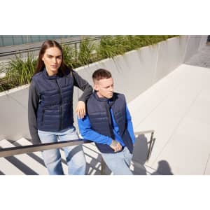 A navy padded vest is worn by a man and woman, posing near a modern staircase surrounded by greenery.