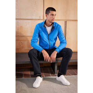 A men's blue blouson jacket displayed on a seated model, paired with dark jeans and white sneakers.