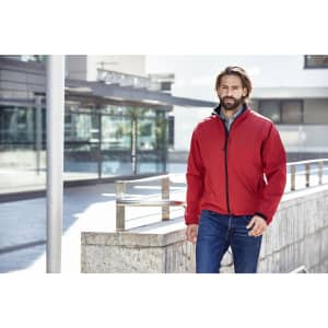 A red men's softshell jacket is displayed on a man walking outdoors near a modern building. The jacket has a logo.