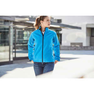 A blue ladies softshell jacket featuring a zip front and a logo, worn by a woman outdoors.