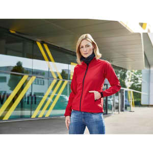 A red ladies softshell jacket with a zip and pockets, displayed on a model against a modern architectural background.