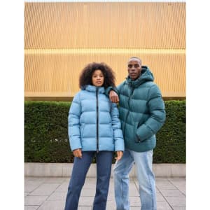 A man in a green puffer jacket and a woman in a blue puffer jacket pose outdoors against a backdrop of greenery.