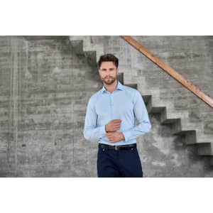 A light blue men's shirt is displayed on a model standing next to a concrete wall and staircase. The shirt has a logo.