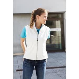 A ladies softshell vest in white, featuring a zip front and a branding logo, styled with a blue top and jeans.