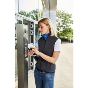 A woman is wearing a grey ladies softshell vest over a blue top, standing outside near a building entrance.