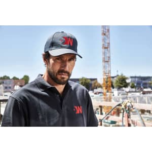 A man wears a black polo shirt and matching cap, featuring a logo, against a construction site backdrop.