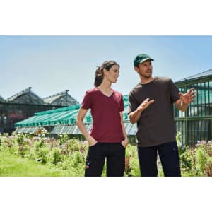 A man in a brown T-shirt and a woman in a burgundy T-shirt stand in a garden setting, wearing workwear.
