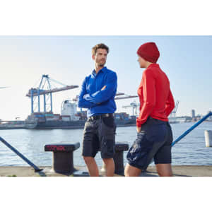 A pair of individuals wearing blue and red work shorts by a waterfront industrial area. The shorts feature pockets and branding.