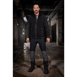 A man stands in a dimly lit industrial space, wearing black and grey workwear pants with a zippered jacket.