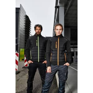A black hybrid jacket for ladies featuring orange accents, worn by a model in an industrial setting.