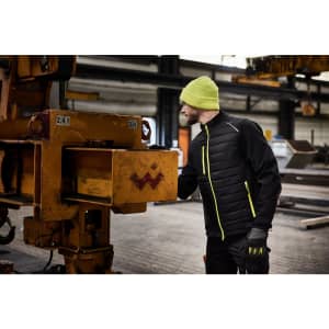 A men's hybrid jacket in black with bright yellow accents, worn by a man in a beanie and gloves near industrial equipment.