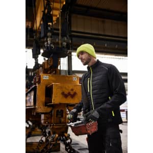 A man wearing a black and yellow hybrid jacket stands in an industrial setting, holding a remote control.