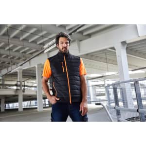 A man in a black hybrid vest with orange accents poses in a spacious indoor setting. The vest features a logo.