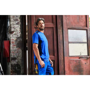A blue men's workwear T-shirt displayed on a model in an industrial setting, featuring a logo.