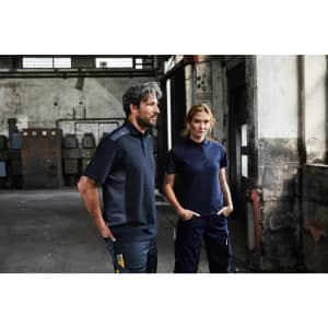 A man and woman stand in an industrial setting, wearing dark blue and navy work polo shirts and matching work pants.