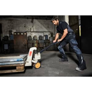A man in dark workwear pants pulls a pallet jack in a warehouse setting. The pants feature pockets and a logo.
