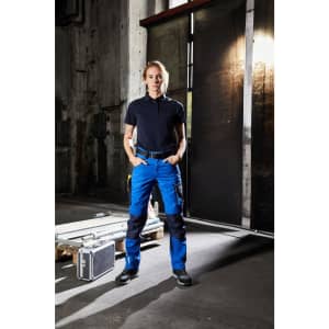 Workwear pants in blue and black, worn by a person standing in an industrial setting. The outfit includes a black polo shirt.