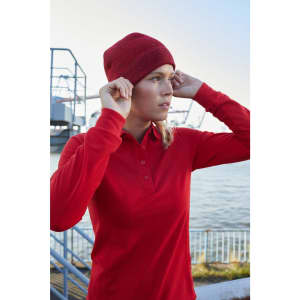 A woman wearing a red long-sleeve polo shirt and a red beanie, standing by the water. The scene features a harbour backdrop.