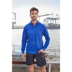 A man stands by the water, wearing a blue long-sleeve polo shirt and black shorts, with a harbour in the background.