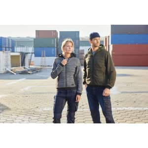 A padded hybrid jacket in grey and black, worn by a woman and a man outdoors near shipping containers. The jackets feature a logo.