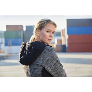 A woman wearing a striped puffer jacket with a black hood, set against a backdrop of shipping containers.