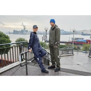 Two models wearing waterproof rain pants in navy and olive green, standing on an outdoor staircase with a waterfront backdrop.