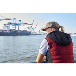 A woman in a red and black padded hybrid vest gazes at a waterfront scene with ships and cranes.