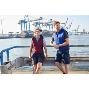 A man and woman wear padded hybrid vests in red and navy, against a backdrop of shipping containers and cranes.