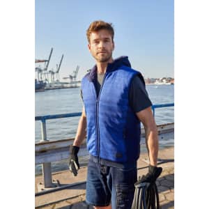 A blue padded hybrid vest with a hood, worn by a man by the waterfront, featuring a logo on the front.