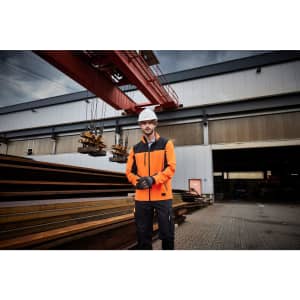 A man in an orange workwear jacket stands outside a warehouse, wearing gloves and a hard hat. The setting includes steel beams and a crane.