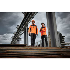 Two individuals wearing bright orange work jackets stand on metal beams at a construction site, with overcast skies in the background.