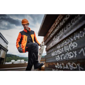 A man wearing an orange and black softshell jacket stands on a ladder beside stacks of metal sheets.
