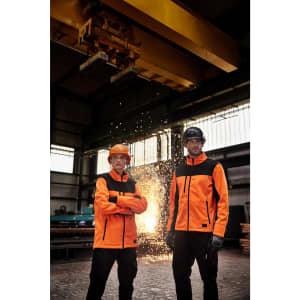 Two individuals wearing bright orange softshell jackets in a workshop setting, with industrial equipment and sparks in the background.