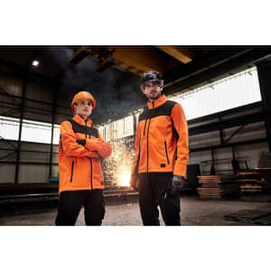 Two individuals wearing bright orange softshell jackets standing in an industrial setting. The jackets have reflective elements and logos.