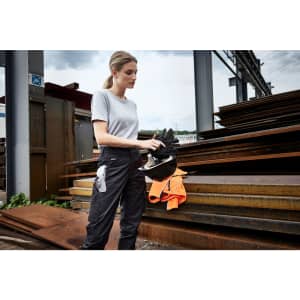 A woman in grey workwear-pants stands holding a helmet, with steel sheets in the background. The pants are dark in colour.