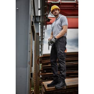 A man in a grey shirt and black work pants stands in front of construction materials, wearing a yellow hard hat and gloves.