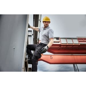A man wearing grey workwear pants and a yellow hard hat climbs a structure. He has gloves on and is on a construction site.