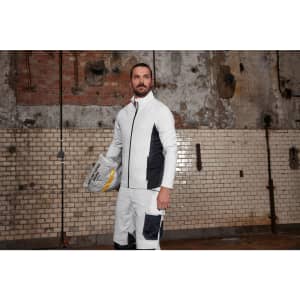 A man wearing a white and black stretch fleece jacket stands in a rustic indoor setting, holding a grey bag.