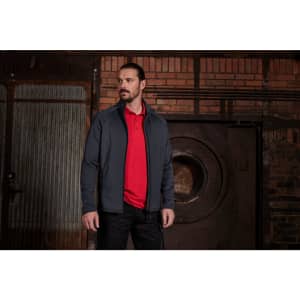A man is wearing a dark grey stretch fleece jacket over a red polo shirt, set against an industrial background.