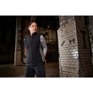A black padded hybrid vest is worn by a model in an industrial setting. The vest has a logo and a sleek design.