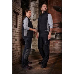 A padded hybrid vest in black, worn by two people, with a grey long-sleeve top underneath. The setting features industrial decor.
