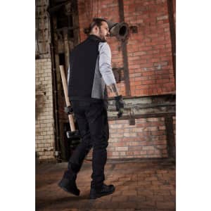 A man wearing a black padded hybrid vest, grey sleeves, black pants, and gloves, walking in an industrial setting.