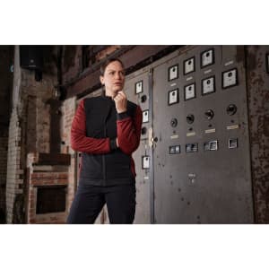 A woman in a black and red padded hybrid jacket stands thoughtfully beside a vintage control panel.